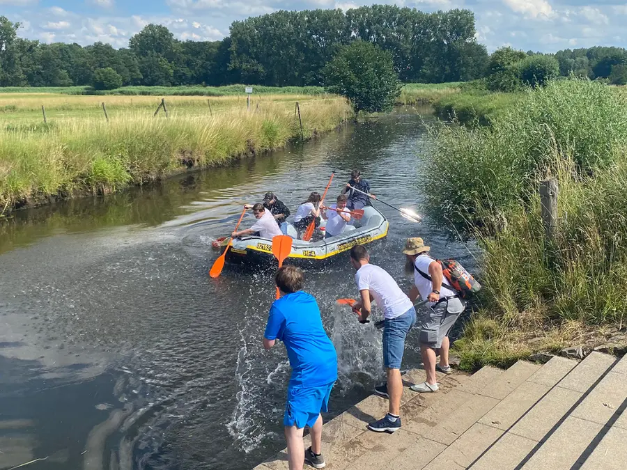 Mitarbeitende paddeln in einem Schlauchboot auf einem Kanal, während andere zusehen – symbolisiert Teamaktivitäten und Spaß.