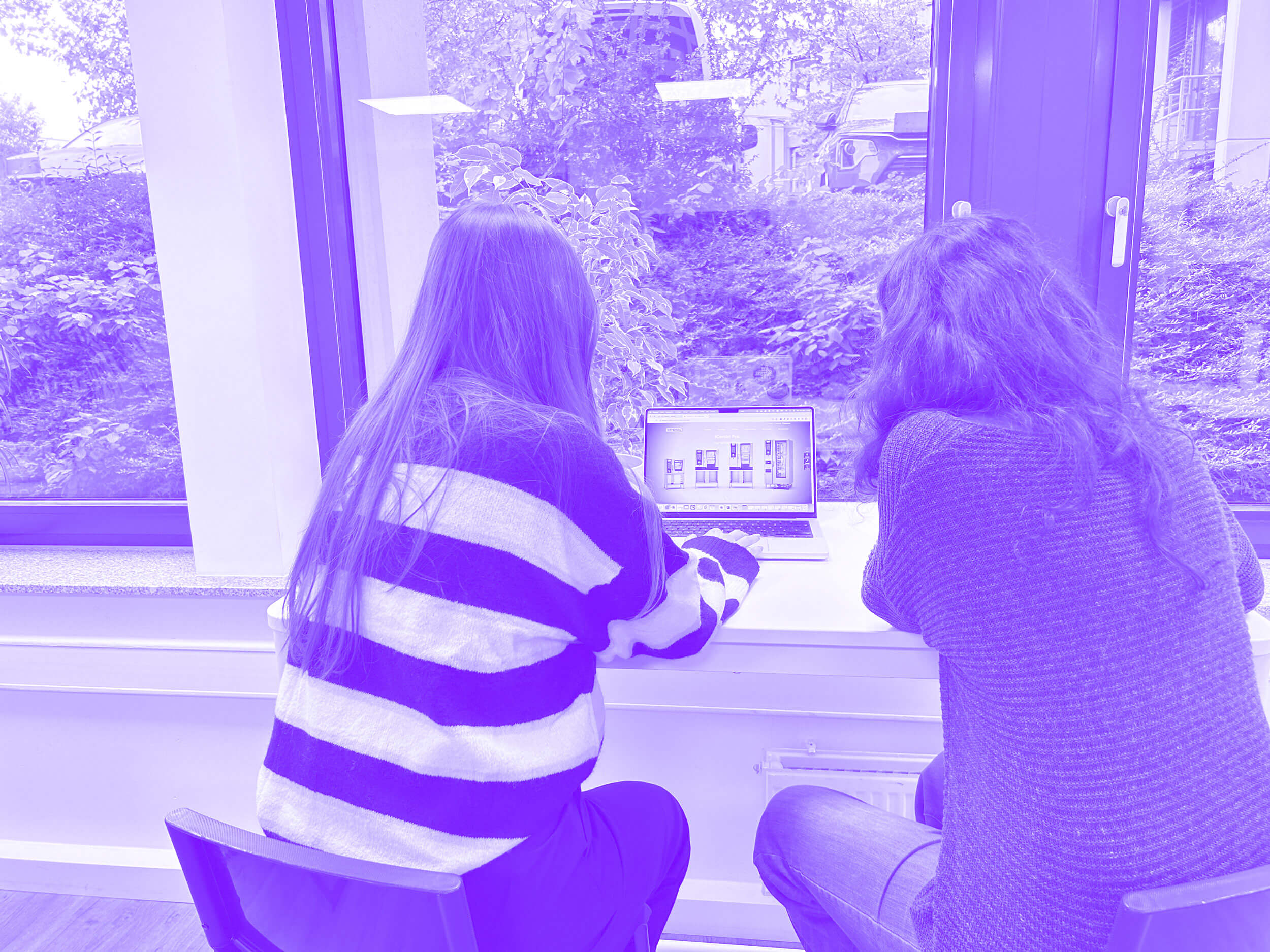 Zwei Mitarbeiterinnen von savotex arbeiten gemeinsam an einem Laptop in einem hellen Raum, mit Blick auf grüne Pflanzen durch große Fenster – symbolisiert Teamarbeit und Produktivität.