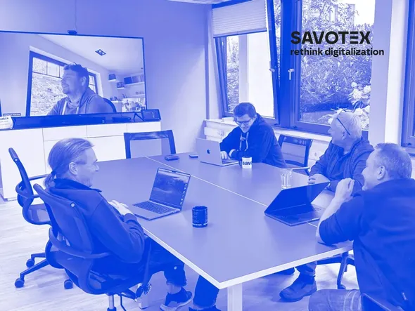 Fünf Gesellschafter der Firma Savotex bei einem Meeting in einem modernen Konferenzraum. Vier von ihnen sitzen mit Laptops um einen Tisch, während der fünfte Gesellschafter per Videokonferenz auf einem großen Bildschirm zugeschaltet ist. Im Hintergrund große Fenster mit Blick ins Grüne. Auf dem Tisch stehen Kaffeetassen, darunter eine mit dem savotex-Logo.