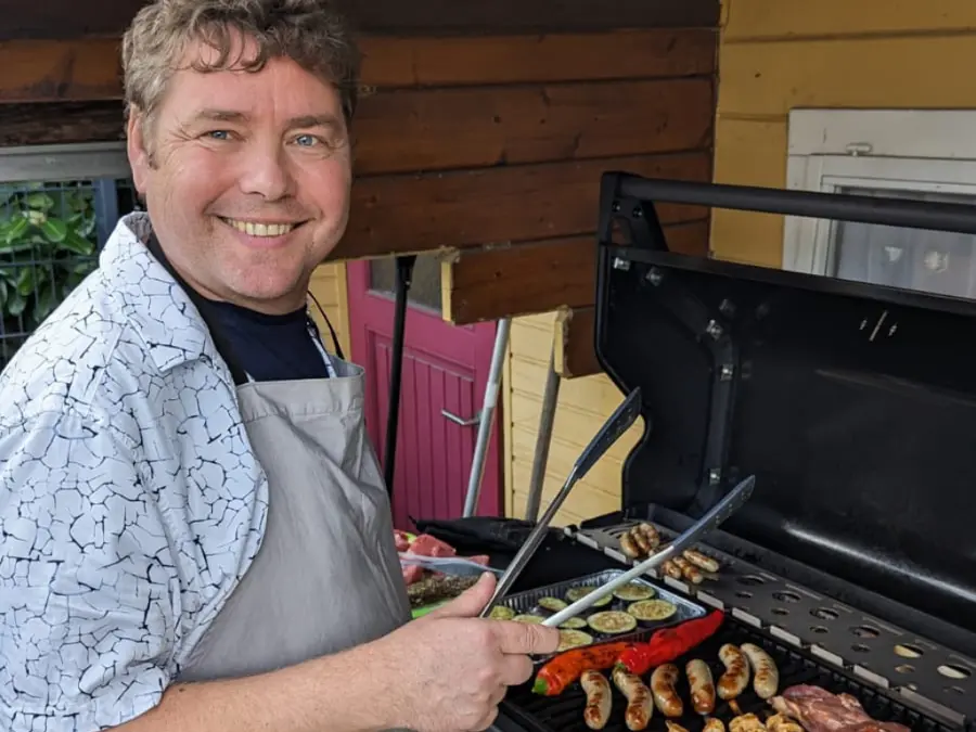 Unser Geschäftsführer Matthias Klautke grillt Würstchen und Gemüse an einem Gasgrill, lächelnd in einer entspannten Umgebung – symbolisiert Team-Events und Freizeitaktivitäten.